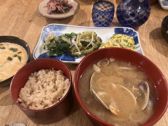 A Japanese meal is presented on a wooden table, featuring a red bowl of miso soup with clams, a bowl of brown rice, a plate with a mix of blanched greens, and a yellow egg dish. In the background, there's  a flask of sake and a sake cup visible