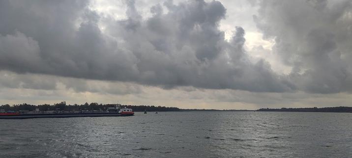 View to the South-West of the Nieuwe Merwede from the local ferry. The lower two-fifths of the photo shows water, dark grey, fairly calm despite the wind. Trees on the horizon, almost black. On the left, just below the horizon, an inland cargo vessel is moving out of the picture, also mostly grey/black, with two red stripes - the only bright colour in the photo. The top three-fifths of the photo consists of grey cumulus clouds, threatening, but without rain.