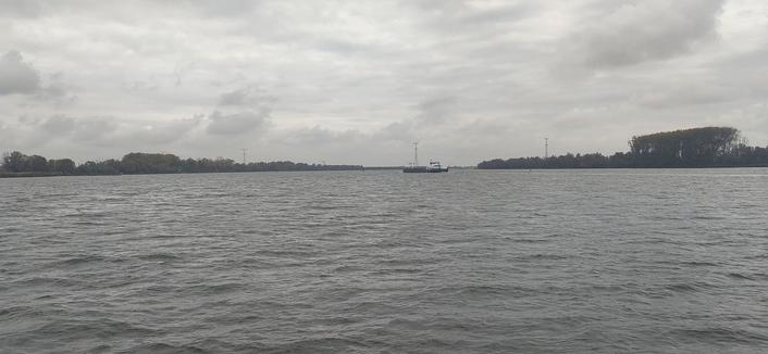 View to the North-East of the Nieuwe Merwede from the local ferry. The lower half of the photo shows water, grey, fairly calm despite the wind. Trees on the horizon, dark grey, some electricity pylons visible. In the middle, just below the horizon in front of one of the pylons, an inland cargo vessel, grey/black, white cabin. The top half of the photo consists of white/grey clouds.