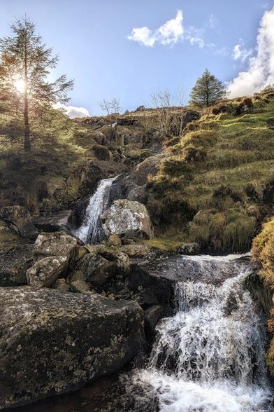 A vertical, outdoor photo of a rocky, multi-tiered waterfall cascading down a steep, grassy hillside under a bright blue sky with scattered white clouds. Two coniferous trees frame the top left and top right, with the sun's lens flare visible behind the left tree. The dark rocks in the foreground are wet and covered in moss. The light brown and green grasses of the hill are illuminated by the strong sunlight from the upper left.