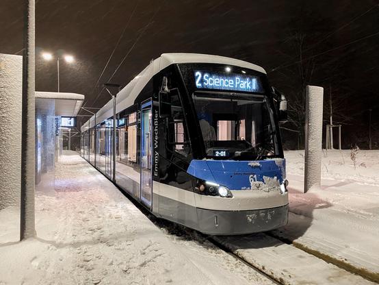 Eine blaue moderne Straßenbahn steht an einer verschneiten Haltestelle. Es ist dunkel und in den Lampen kann man sehen dass es stark schneit. Am Zielanzeiger der Tram steht Science Park II