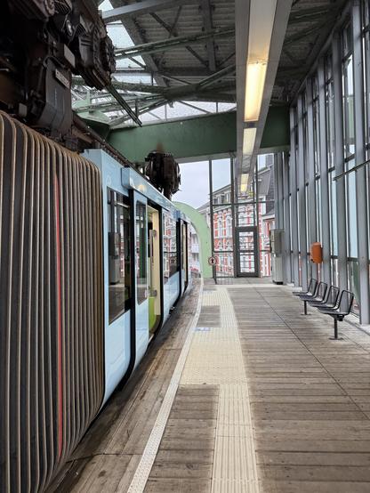 Schweebahnzug im Bahnhof Vohwinkel. Links ist der Zug mit geöffneten Türen. Das Bild ist auf Höhe des vorderen Knickgelenks aufgenommen. Rechts ist der Bahnsteig aus Holz. Der Zug hängt ca. 15 cm oberhalb des Bahnsteigs.