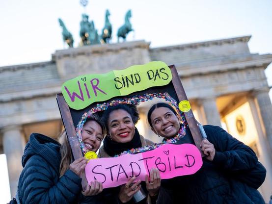 Ein Gruppenfoto von drei lächelnden Frauen, die ein großes, rechteckiges Schild in Form eines Bilderrahmen-Ausschnitts halten, durch den ihre Gesichter sichtbar sind. Das Schild trägt die Aufschrift „WIR SIND DAS STADTBILD“ in handschriftlicher Form. Die Wörter „WIR SIND DAS“ stehen auf dem oberen, neongrünen Teil des Rahmens, und das Wort „STADTBILD“ steht auf dem unteren, pinkfarbenen Teil, der mit buntem Konfetti-artigen Material umrandet ist. Die Frauen stehen draußen, im Hintergrund ist das Brandeburger Tor in Berlin zu sehen, beleuchtet von der Abendsonne.

(Beschreibung von Gemini)