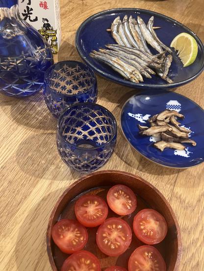 A wooden table displays a selection of Japanese cuisine items: a plate of small, dried fish garnished with a slice of lemon, slices of fresh tomatoes in a round wooden bowl, and a small dish with dried mushrooms. There are also two intricately patterned sake glasses on a wooden table top