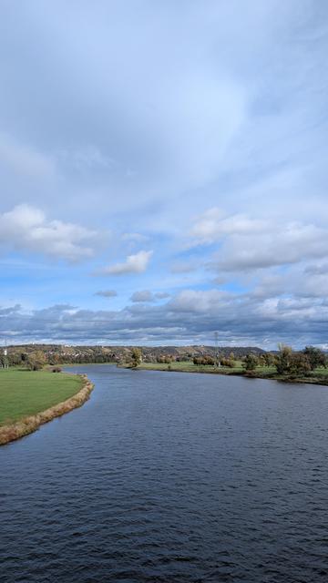 Blick von der Elbbrücke Niederwartha auf die Elbe. Der Himmel ist bewölkt, aber die Sonne findet ein paar Lücken. Links und rechts der Elbe sind ausladende Wiesen mit vereinzelten Bäumen. Es ist leicht windig und die Elbe etwas aufgewühlt.