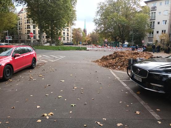 Fast 2 meter hoher Laubberg, der in einer Rechtskurve einer engen 2Richtungsstraße von einer Sperrfläche über die weiße Linie auf die Fahrbahn abgerutscht ist. Der Kreisverkehr im Hintergrund ist halb gesperrt, sämtliche Fahrzruge müssen hier  sehr scharf, mehr als 270 Grad, rechts um die Ecke fahren. Unter dem Laubberg vergraben ist auch eine nicht mehr  sichtbare und jetzt unbenutzbare Fußyquerung