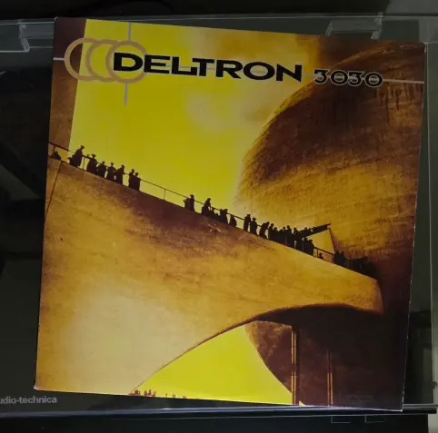 The image shows a large, abstract sculpture in a museum or gallery setting. The sculpture is predominantly white with a rounded, domed shape at the top, resembling a large, inverted bowl. Several figures of people are gathered around and beneath the sculpture, some looking up at it. The background is blurred and indistinct, suggesting depth and space within the exhibition area. "DELTRON" is written in large, bold black capital letters on the left side of the image.  "30" is written in large, bold black capital letters on the right side of the image. "studio-technica" is written in small letters at the lower right of the image.