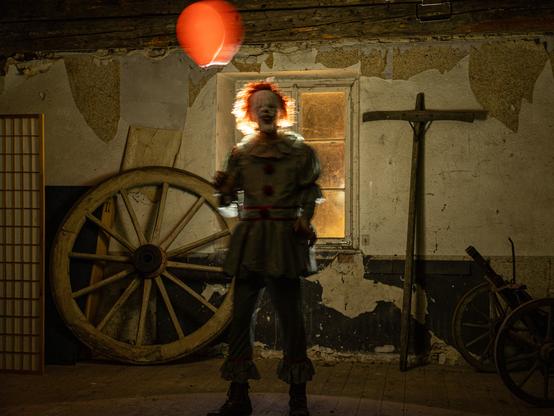 Ein Clown mit einem roten Ballon steht vor einem Fenster in einem dunklen Raum. Die Tapete an der Wand blättert ab und überall hängen Spinnweben. Ein kreuzförmiges Stück Holz und ein großes Wagenrad lehnen an der Wand. Der Clown wird von hinten beleuchtet und sein rotes Haar leuchtet. Das Bild des Clowns ist unscharf, weil er sich bewegt. 

A clown with a red balloon stands in front of a window in a dark room. The wallpaper on the wall is peeling and cobwebs hang everywhere. A cross-shaped piece of wood and a large wagon wheel lean against the wall. The clown is lit from behind and his red hair glows. The clown’s image is blurred because he is moving.
