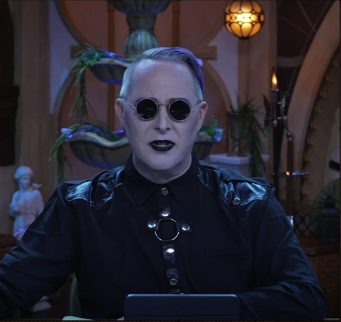 A screenshot from Critical Role
Taliesin Jaffe is dressed in a severe outfit, solid black, with black leather straps and metal buckles and rings and clasps.
His hair is shaved close on the sides, with a sharp part to one side and the rest combed over and bright purple.
On his face are black lensed round rim glasses perfectly set in his eyes, and dark black lipstick. His skin is an ashen grey.
He looks very much not amused.