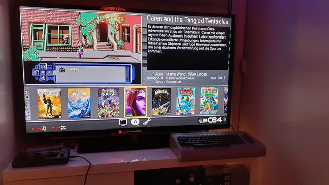 Man sieht das Spielekarussel (das Spielemenü) vom TheC64 Mini auf einem schwarzen LCD-TV. Der TV steht auf einer weißen TV-Bank vor eine weißen Wand aus Holzpanelen. Links vorm TV liegt der TheC64 Mini Black Edition und rechts vorm TV liegt der TheC64 Maxi.