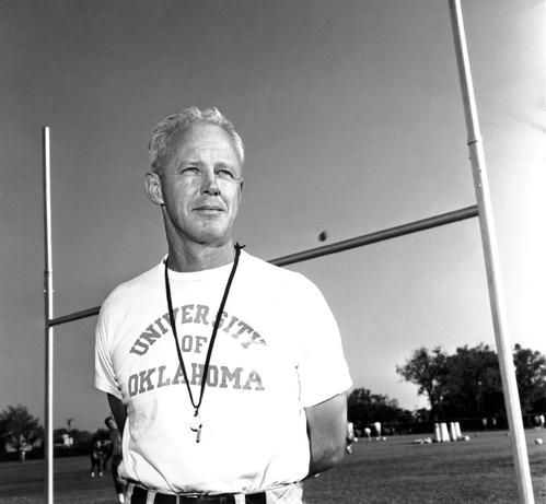 A black and white photo of Bud Wilkinson wearing a shirt that says University of Oklahoma on a football field in front of a goalpost