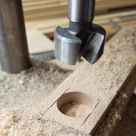 Zu sehen ist ein Stück Ahornholz mit einem Loch und Resten einer schwarzen Markierung, umgeben von vielen Holzspänen. Am oberen Bildrand ist ein großer Holzbohrer abgebildet.