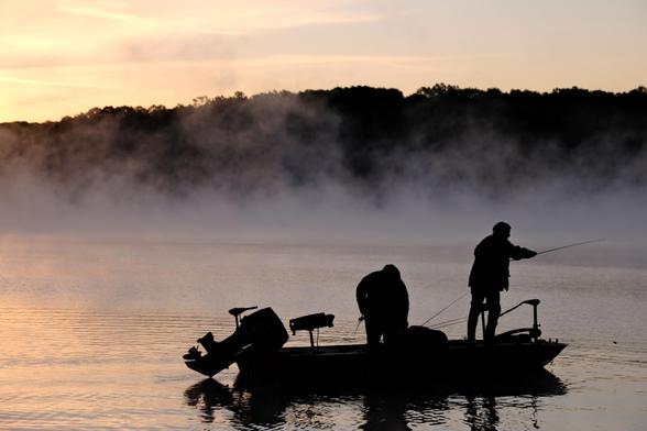 Two fishermen on a boat on a lake, as fog rises from the water. Predawn light is reflected on the water.