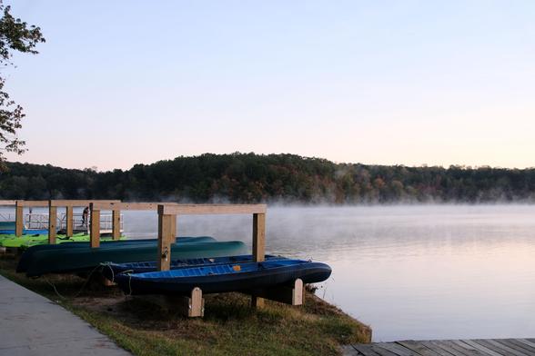 A lakeview before sunrise as fog rises from the water. Blue and green kayaks on a wooden rack in the front.