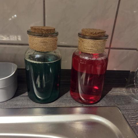 Zwei Glasflaschen mit rotem und grünem Spülmittel darin, welche aussehen wie Zaubertrank-Flaschen aus einem Videospiel.