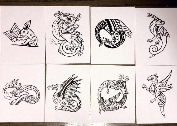Eight sheets of white paper with black block prints on them, they are: a bunny reading, a filigree wyvern, a grumpy looking wyvern lik creture forming a circle with its head/neck and tail, a horned, fancy and stuck up looking wyvern with a long curled tail, a happy smiling wyvern like creature looking over its shoulder, an offended looking wyvern with wings extended and and flames coming from its mouth with a a second smaller and astonished head on its tail, an oroboros made of two wyverns biting each others tails, one black and one white, and a small, dainty, and concerned looking wyvern with feathery wings and a curled tail flying with one paw extended as if to pat someone on the shoulder