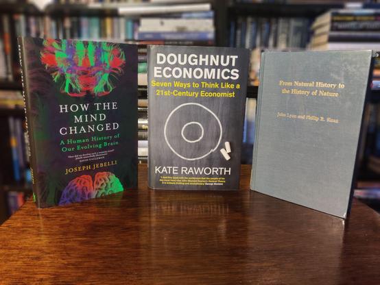 A photo of three books standing on a small, brown, wooden table. The out-of-focus background shows black shelves full of books.

On the left, standing up, How the Mind Changed, showing two pictures of a human brain on a black background. At the top, part of a connectome picture, showing a high-resolution brain scan of neuronal connections in green, red, and purple; at the bottom, part of the surface of the brain, using the same green, red, and purple colour scheme 

In the middle, Doughtnut Economics, showing a close-up picture of a blackboard with a drawing in white chalk of two concentric circles, one inside the other. Two pieces of white chalk lie next to the outer circle.

On the right, From Natural History to the History of Nature, showing a plain hardcover book bound in navy blue cloth with gold lettering. This is an ex-library copy bound in transparent protective foil that gives it a slightly shiny look.