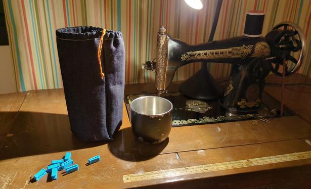 An old zenith treadle sewing machine desk have a stainless steel cup and jean coloured pouch sewn for it side by side. There is a colourful striped curtain in the background.