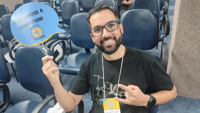 Foto de um rapaz bonito, de barba, óculos e camisa preta sorrindo e segurando uma placa que diz "Code, coffee, connection".