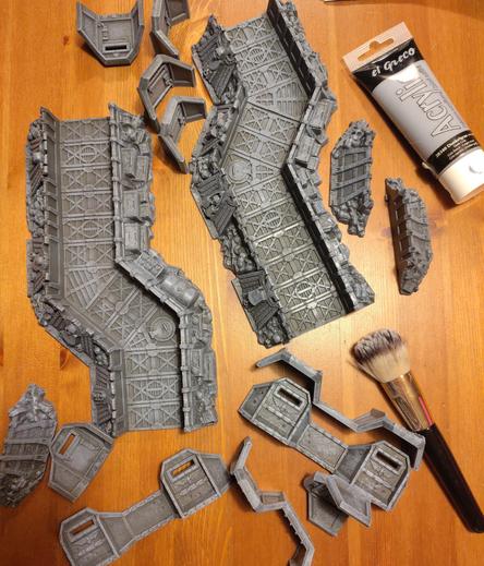 A load of terrain pieces on a table - they are dark grey with a drybrush effect in a lighter effect, accenting all edges and raised surfaces, giving more depth to the structures and textures.
Next to them is a plastic squeeze tube of acrylic paint and a large cosmetic brush.