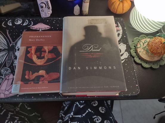 On my desk are pumpkins and mushrooms, all in green, orange, and pink shades. Resting on the desktop is a pair of books. One is Mary Shelley's Frankenstein with an orange Barnes and Nobles' Classics cover, with art of the human skull on it. Next to it is Drood from Dan Simmons. On the cover is the silhouette back of a dark figure with a top hat and coat.