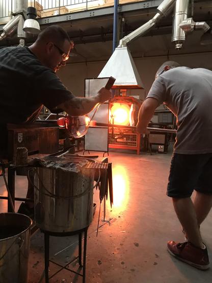 Deux hommes techniciens verriers travaillent sur une pièce en verre dans l'atelier du CIRVA, à l'arrière plan on aperçoit de la lumière qui émane d'un four ouvert.