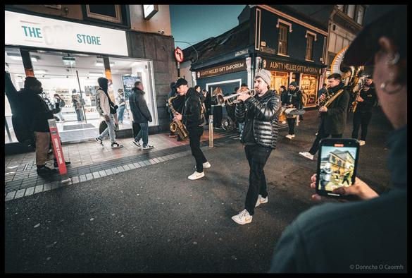 Brass band musicians performing on Patrick Street during Cork Jazz Festival parade at dusk, with trumpeter wearing sunglasses and puffer jacket in centre foreground, surrounded by crowd of spectators many filming on mobile phones, with brightly lit Penneys store and other Patrick Street shops visible in the background under evening sky.