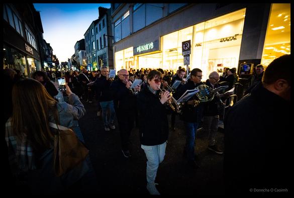Brass band musicians marching down a Cork street during Jazz Festival parade at dusk, with trumpet player in beanie and puffer jacket in foreground, spectators watching from shop doorways, and someone filming on mobile phone visible in the right foreground, with turquoise evening sky above.