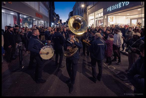 Lamarotte brass band performing on Patrick Street during Cork Jazz Festival parade at dusk, with massive sousaphone bell dominating the frame, drummer visible on left, surrounded by large crowd of spectators outside Guineys and Penneys stores under evening sky.