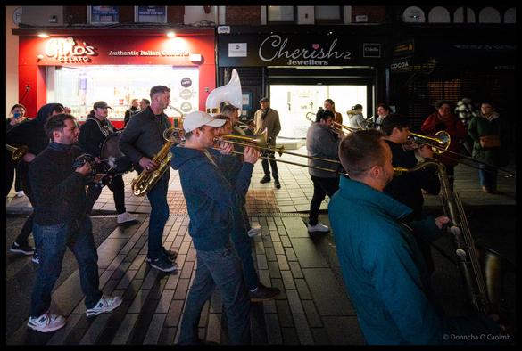Brass band musicians performing on Patrick Street in Cork during the Jazz Festival parade at night, with trombonists and saxophonists in casual clothing playing energetically outside Gino's Italian restaurant and Cherish jewellers, with crowd of spectators visible in the background.