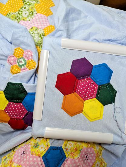 Light blue pillow case, partially in a rectangle quilt hoop. There are colourfull applications on the cloth. In the center is a rainbow flower with quilting in process.