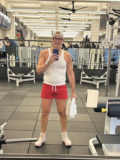 A man (me) is taking a selfie in a gym. He is wearing a white tank top, red shorts, and light pink training shoes. He holds a towel in one hand and his phone in the other. The gym background features various exercise equipment and mirrors.