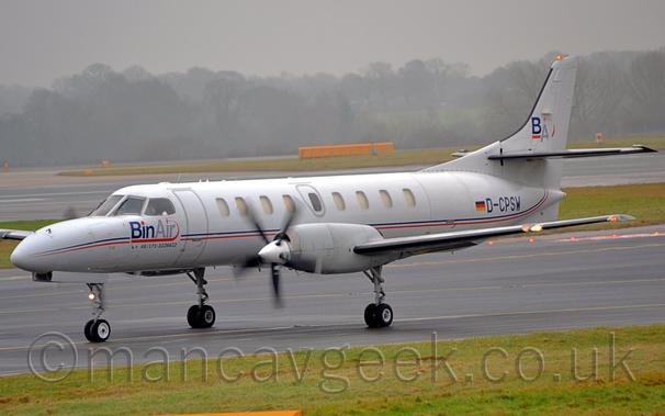 Side view of a twin-engined jet airliner taxiing from right to left.
The plane is basically a long, thin tube with wings, closed off at both ends.
It is mostly white, with a thin blue and red stripe running along the body, with blue and grey "BinAir" titles under the cockpit window, and the black registration D-CPSW next to a small German flag on the rear fuselage.
There is a blue and grey "BA" logo on the tail, underlined in red.
Green grass fills the foreground, with more grass in the background separating this taxiway from a runway, then leading off to trees slowly vanishing in to hazy grey clouds in the distance.
