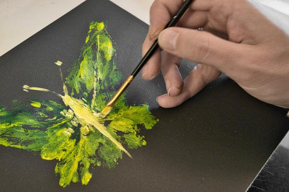 Eine Nahaufnahme zeigt die Hand einer Person, die mit einem Pinsel vorsichtig Details auf einem leuchtend grünen und gelben Schmetterling auf schwarzem Papier malt.

Die Komposition ist nah und fokussiert, wobei die Szene im oberen rechten Bereich von einer Hand dominiert wird, die einen feinen Pinsel hält, der gerade eine der Flügel des Schmetterlings berührt. Der Schmetterling nimmt den größten Teil der Bildmitte ein und scheint vor dem dunklen Hintergrund aufzutauchen. Der Pinsel ist im Begriff, einen Teil des Flügels zu bemalen, was das Engagement des Betrachters in den kreativen Prozess vertieft. Das Bild wird von links oben beleuchtet, wodurch ein subtiler Schatten auf dem Papier entsteht und die Textur der Oberfläche verstärkt wird.

Der Hauptgegenstand ist ein Schmetterling, der mit grünen und gelben Farbtönen gemalt wurde. Die Flügel werden mit verschiedenen Grüntönen und einigen gelben Akzenten dargestellt, die eine lebendige und fast fluoreszierende Wirkung erzeugen. Die Details sind noch nicht vollständig ausgemalt, wodurch ein Gefühl der Unvollkommenheit entsteht, das die Kreativität des Künstlers und den laufenden Malprozess hervorhebt. Die Hand, die den Pinsel hält, ist sanft und konzentriert, mit sichtbaren Details wie der Hautstruktur und kleinen Farbspritzern, die auf die Aktivität hinweisen.

Die verwendeten Medien sind Acryl- oder Ölfarben, die auf schwarzem Papier aufgetragen werden. Die Verwendung von dunklem Papier dient als Kontrast für die leuchtenden