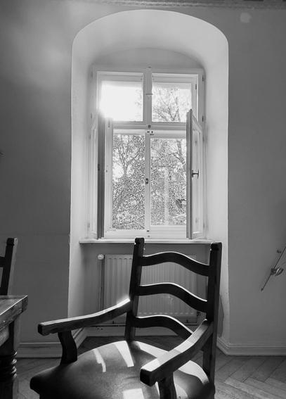 Ein Schwarzweißfoto eines alten gepolsterten Lehnstuhls aus dunklem Holz mit Armlehnen und drei geschwungenem Querstreben in der Rückenlehne steht vor einem geöffneten Fenster, durch das die Sonne in den Raum scheint. Die Wand rund um das Fenster ist weiß gestrichen.

An old upholstered armchair made of dark wood with armrests and three curved crossbars in the backrest stands in front of an open window through which the sun shines into the room. The wall around the window is painted white.