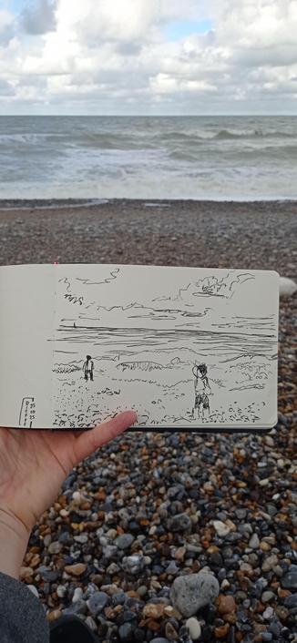 Une main tient un carnet de croquis devant la mer et une plage de galets. Sur le carnet un croquis à l'encre représentant le même paysage + des silhouettes d'enfants.
