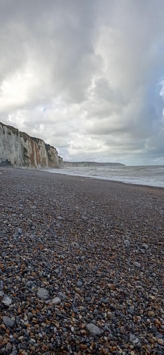 Photo de plage  de galets avec falaise en fond, ciel nuageux mais lumineux.