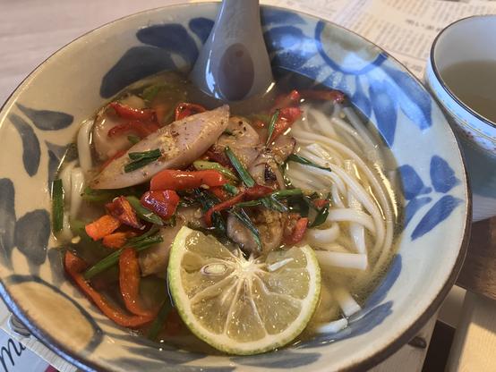 A bowl of noodle soup featuring thick noodles, slices of fish sausage, and colorful vegetables, garnished with fresh herbs and a slice of lime. A spoon rests in the bowl, with a teacup visible in the background.