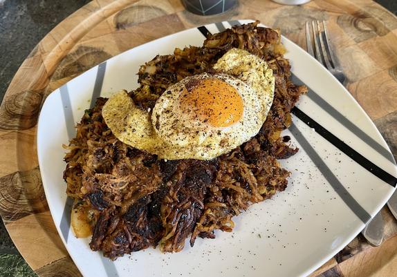 a square plate on a round butcher block board. a fork is visible to the right. on the plate is a mess of crispy brown hash, with shreds of meat and noodles visible. on top is a fried egg. it’s all dressed with herbs and spices.