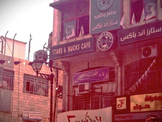 Old pic of Stars & Bucks Cafe buidling entrance. Logo is green like Starbucks, the name is also written in arabic, we see a light beige brick building on the left, in classic Palestine style.