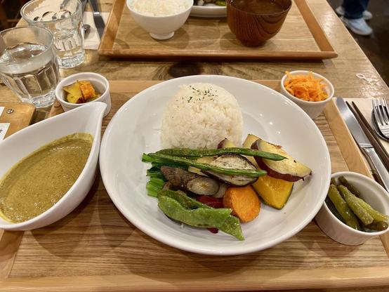 A plate of rice accompanied by a variety of colorful perfectly grilled vegetables, including green beans, carrots, burdock, sweet potatoes and kabocha squash, along with a small bowl of coconut curry. Additional side dishes include pickled vegetables and green beans. They are on a wooden tray on the wooden table. There are two glasses of hot water on the table. Also another tray of Japanese set of dishes on the other side of the table, which my friend had.