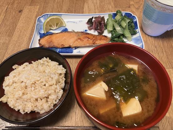 A traditional Japanese meal featuring a bowl of brown rice, a bowl of miso soup with tofu and seaweed, grilled salmon garnished with a slice of lime, and a side dish of pickled vegetables and sautéed greens on a plate. A