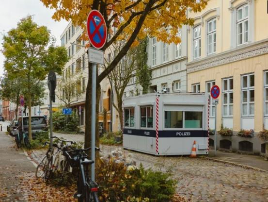 Schmale Wohnstraße mit mehrstöckigen Häuser (Altbauten). Mehrere Bäume am Straßenrand. Dazu ein kleiner Baucontainer mit der Aufschrift "Polizei"