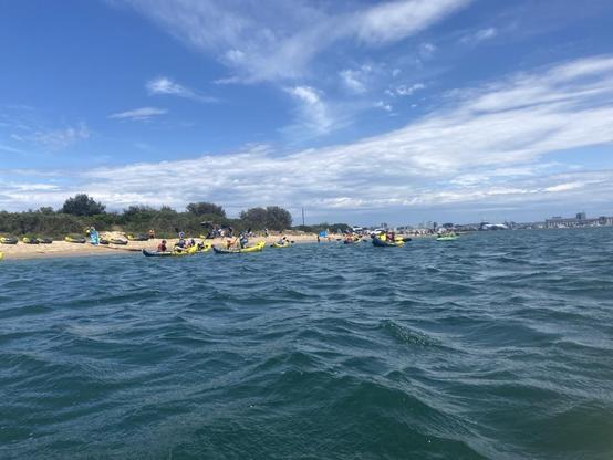 Looking towards shore at Horseshoe Beach with people in bright yellow and grey inflatable kayaks in the water and people up on the sandy beach. Sunny day, light layer of cloud across the blue.