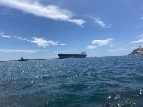 A big coal ship coming in to Newcastle harbour with Nobbys to the right and a tug boat to its left. We're in the water kayaking when i took the shot. The black end of the paddle is visible bottom right.