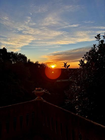 A vibrant sunrise over a landscape, framed by silhouettes of trees. In the foreground, a balcony with a decorative pot containing flowers is visible, adding depth to the scene. The sky features colorful hues of orange and blue, with scattered clouds.
