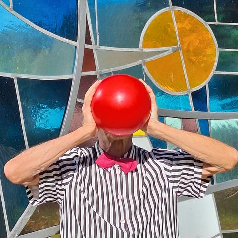 Ein Mann in hohem Alter, bekleidet mit einem kurzärmeligen, vertikal schwarz-weiß gestreiftem Hemd und magentafarbener Fliege hält mit beiden Händen einen roten Ball vor sein Gesicht.
Er steht vor einer Bleiglasscheibe, die sich aus stilvollen Glasblöcken, kunstvollen Mosaiken und abstrakten Farbflächen zusammensetzt.
Rechts oberhalb vom Ball ist ein kreisrundes, orange-leuchtendes Element in das von links kommend die Spitze eines sichelförmigen Elements hineinragt.
Die Farbgebung der Mosaikteile in Grau, Braun und Blau wird durch die Beschaffenheit des Hintergrund beeinflusst.
Die Sonne scheint und gibt dem Bild eine besondere Strahlkraft.

An elderly man, wearing a short-sleeved, vertically striped black and white shirt and a magenta bow tie, holds a red ball in front of his face with both hands. He stands in front of a leaded glass panel composed of stylish glass blocks, artistic mosaics, and abstract colored areas. To the right above the ball is a circular, glowing orange element, into which the tip of a crescent-shaped element protrudes from the left. The gray, brown, and blue color scheme of the mosaic pieces is influenced by the nature of the background. The sun is shining, giving the image a special radiance.