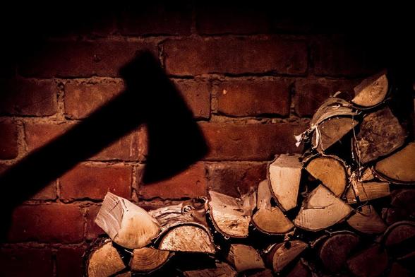 Shadow of an axe positioned over a stack of chopped wood logs against a textured red brick wall. The lighting is dim, creating a dramatic contrast between the shadow and the background. The logs are unevenly stacked, displaying various cuts and textures.