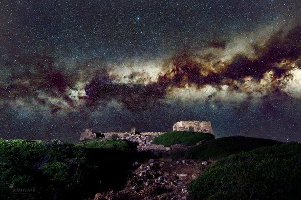 A composite photo of a night scene of a ruin under the stars and Milky Way. The ruins sit on top of a hill with green shrubs, and the Milky Way spans from left to right in the night sky. Many stars are visible, like diamonds in the sky.
