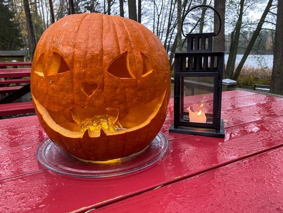 Kahvilan terassin punaisella, märällä puupöydällä on kurpitsalyhty ja tavallinen kynttilälyhty. Kurpitsan suussa led-valoketju.